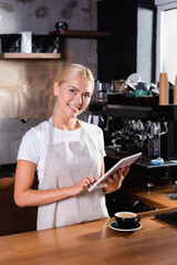 Blonde barista using digital tablet near cup of coffee on bar counter