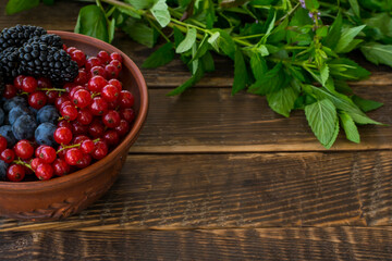 Fresh summer berries in a clay bowl on a wooden table. In the frame there is a part of a bowl with red currants, blueberries and blackberries, in the background mint branches. Copy space. Summer food