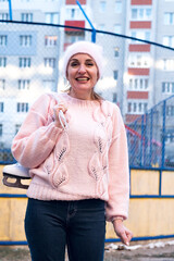 Beautiful woman weared in pink sweater and hat with ice skates on the back walks in winter snowy...