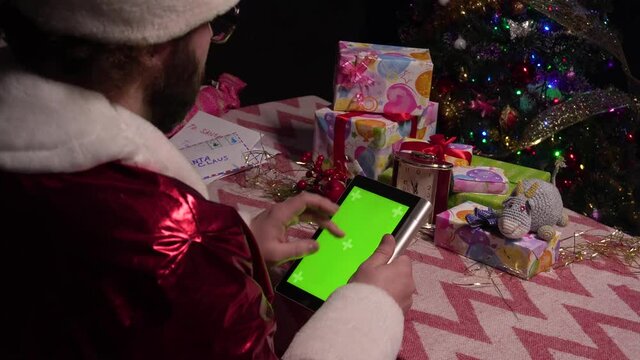 Santa Claus is using the green screen of the tablet for shopping.Santa Claus sits at a table with a tablet in his hands and flips through web pages. Tablet with a green screen. chroma key.