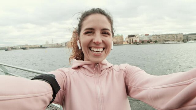 Medium shot of beautiful Caucasian sportswoman wearing sport clothes and wireless headphones standing on embankment during a run and shooting herself on video and laughing