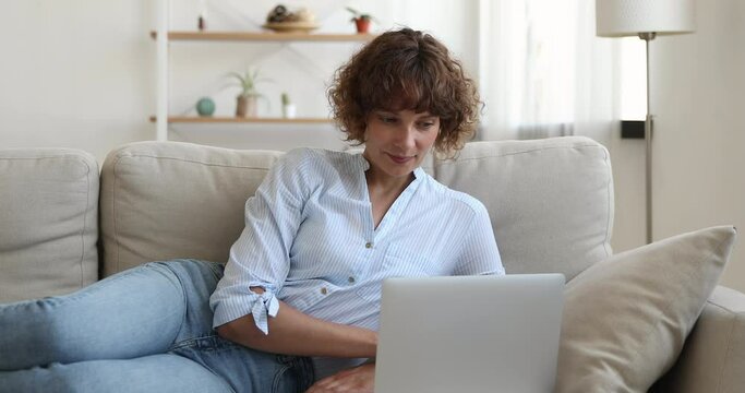 Happy middle aged relaxed woman lying on comfortable couch, spending free weekend leisure time communicating with friends in social networks, shopping online on computer, watching funny photos videos.