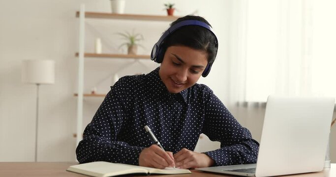 Beautiful Indian woman sit at desk using laptop listen audio course through wireless headphones makes notes on copybook enjoy language learning, develop knowledge. Self-education, e-learning concept