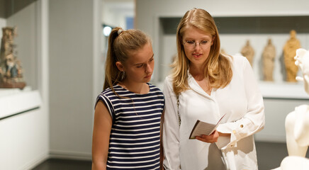 Young woman visitor with daughter with guide book looking at exhibition in museum of art