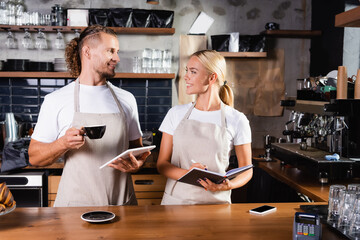  barista with coffee cup and digital tablet near waiter with notebook, and payment terminal on bar...