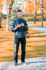 Youthful Satisfied Guy Using Black Smartphone at the Beautiful Autumn Park. Handsome Smiling Young Man with Mobile Phone at Sunny Day - Long Shot Portrait