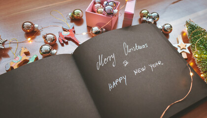 A black paper notebook with Merry Christmas and Happy New Year in silver letters surrounded with colorful Christmas decorations, ornaments and lights on wooden background. Happy holidays background. 