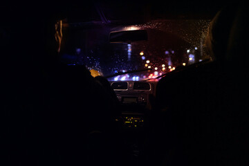 car drives through the night city in the rain, blurry lights of street lamps and traffic lights on the street, the window is torn by water drops, traffic concept