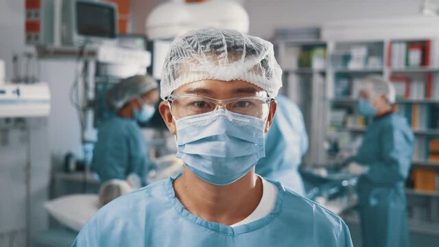 Saving lives. Portrait of the young asian doctor wearing medical face mask and looking at the camera.Medical worker standing at the medical ward with doctors at the background. Concept of safety lives