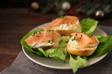 Baguette canapes with salmon and cream cheese on a plate with lettuce, dark rustic wooden table with Christmas decorations, copy space