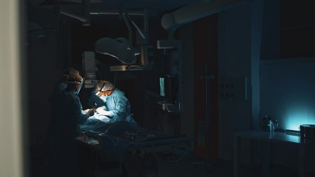 View from the hall of the professional doctors wearing blue medical clothes in the dark operation theatre under the bright light of a surgical lamp doing operation for patient at the serious condition