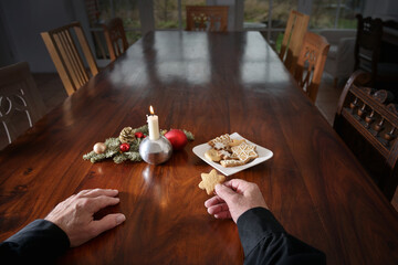 Hands of a lonely old man with a cookie sitting at a big family table with candle, Christmas...