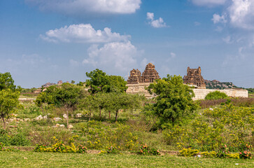 Fototapeta na wymiar Hampi, Karnataka, India - November 4, 2013: Seen from Queens bath structure of Vijayanagara Empire are ruins of Chandrashekhara Temple behind green belt and under blue cloudscape.