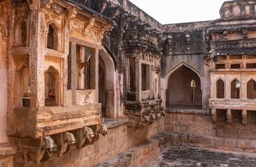 Naklejka premium Hampi, Karnataka, India - November 4, 2013: Queens bath structure of Vijayanagara Empire. 3 brown stone balconies over dry pool in corner of structure. Black mold and silver sky.