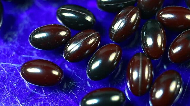 Maroon and black oval spindle-shaped capsules in a gel shiny shell extremely close-up moves from bottom to top and from top to bottom. Real time, natural light, on a blue background, carotenoids, caro