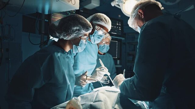 Skillful team. Group of smart skilled surgeon wearing protective gear standing together near the patient, holding medical clamps and helping each other while working in the team. Teamwork of doctors