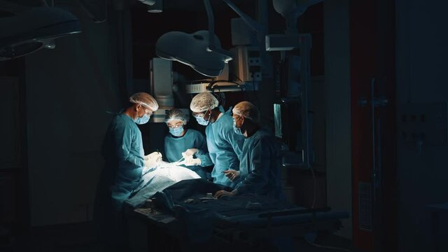 View from the doors at serious surgeon team performing cardiothoracic surgery in surgery dark room with medical stuff. Patient lying on table under glowing lamp in hospital with life support system