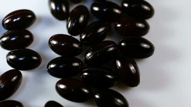 Black oval spindle-shaped capsules in a shiny gel shell pour down. Real time, natural light, white background, carotenoids, carotenoid pigments, Astaxanthin
