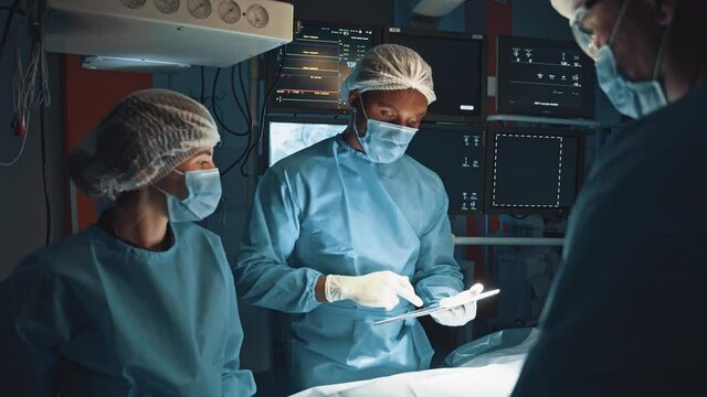 Healthcare and modern medicine. Serious multirace doctor looking together with his team at X-ray and CT results showing at digital tablet while preparing at the serious operation at operation room