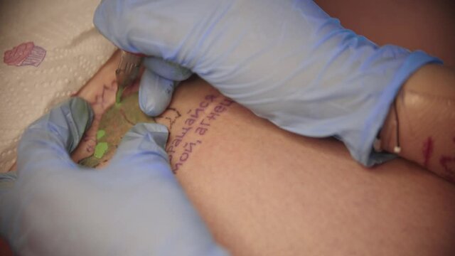 A woman getting a green dinosaur tattoo with russian words in blue ink
