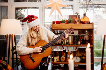 Beautiful young blonde woman in a funny red beanie Santa Claus plays the guitar.