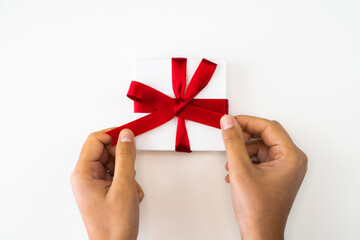 Beautiful holiday or Christmas background image close up of hands from a mixed race African American woman tying a bow with red ribbon onto a small white gift box with white backdrop and copy space.