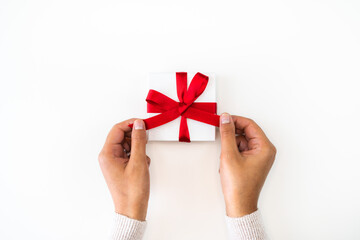 Holiday or Christmas background or backdrop image close up of hands from a mixed race African American woman wearing a sweater tying a red ribbon bow onto a small white gift box with copy space.