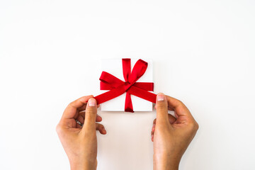 Beautiful holiday or Christmas background image close up of hands from a mixed race African American woman tying a bow with red ribbon onto a small white gift box with white backdrop and copy space.