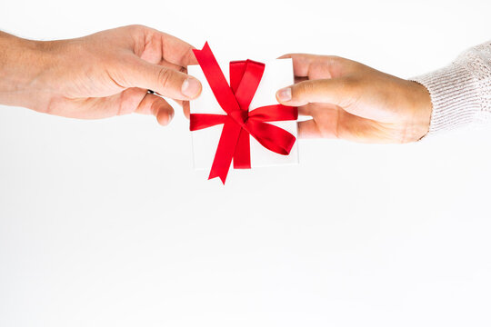 Beautiful holiday or Christmas background image of the hands of a Caucasian man and mixed race woman exchanging a small red ribbon wrapped white gift box on a white backdrop and copy space.