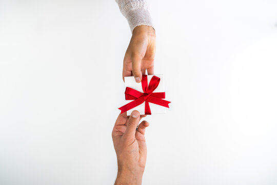 Beautiful holiday or Christmas background image of the hands of a Caucasian man and mixed race woman exchanging a small red ribbon wrapped white gift box on a white backdrop and copy space.
