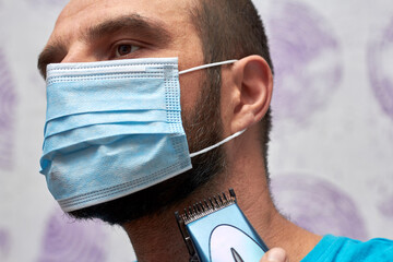A bearded man with a medical mask on his face tries to shave with a trimmer.