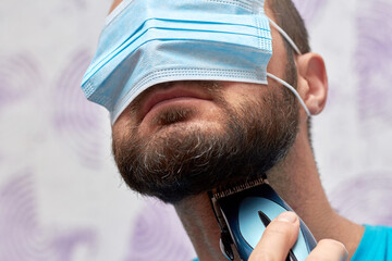 A bearded man with a medical mask on his face tries to shave with a trimmer. Hygiene and the virus...