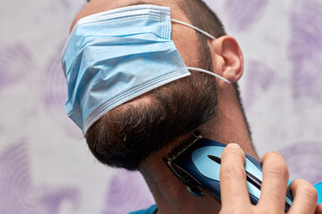 A bearded man with a medical mask on his face tries to shave with a trimmer. Hygiene and the virus...