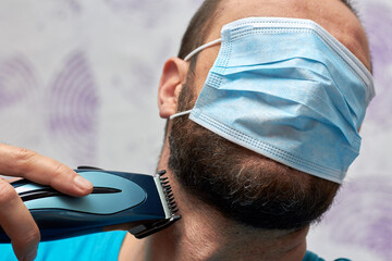 A bearded man with a medical mask on his face tries to shave with a trimmer. Hygiene and virus.