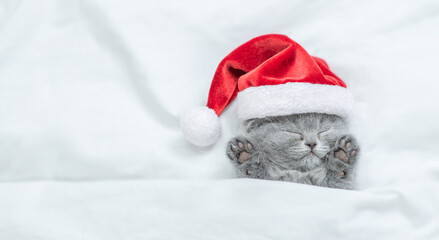 Funny kitten wearing red santa's hat sleeps under a white blanket on a bed. Top down view. Empty...