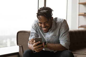 Overjoyed millennial African American man look at cellphone screen have fun using gadget. Smiling...