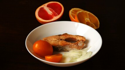 chum salmon steak with onion and yellow tomato salad in a white plate with high sides in a composition with brightly cut orange and grapefruit on a dark rich brown wooden surface