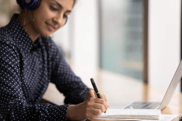 Crop close up of young happy Indian woman study distant on laptop, handwrite in notebook....
