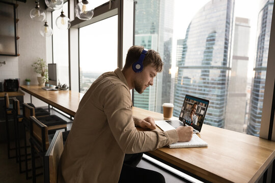 Focused young Caucasian man in headphones sit at desk make notes study distant talk on video call on laptop. Millennial male in earphones have webcam digital virtual conference or event on computer.