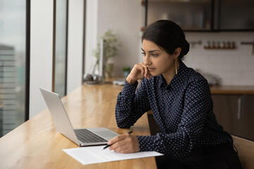 Thoughtful young Indian woman look at laptop screen working online in office. Pensive millennial ethnic female employee use computer watch webinar or training on gadget. Education concept.