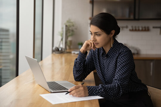 Thoughtful Young Indian Woman Look At Laptop Screen Working Online In Office. Pensive Millennial Ethnic Female Employee Use Computer Watch Webinar Or Training On Gadget. Education Concept.