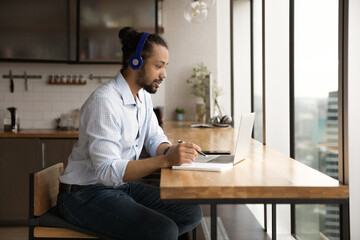 Millennial African American male employee in headphones look at laptop screen working online on...