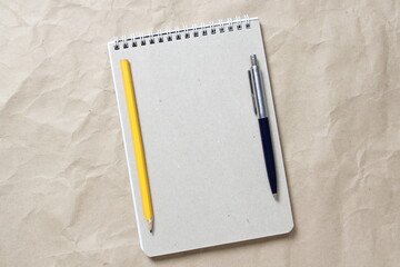  Gray notepad with white coiled spring and pen and pencil on a background of beige crumpled craft paper