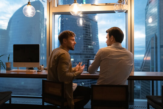 Back view of businesspeople work in office late hours, discuss company business project on computer together. Male colleagues coworkers busy at meeting at workplace at night or evening.
