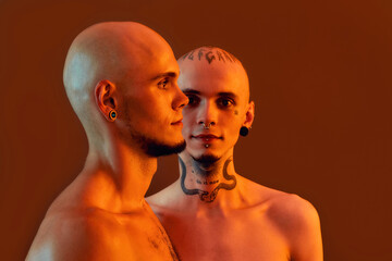 Front and side view of young half naked twin brothers with tattoos and piercings posing together, standing isolated over orange background