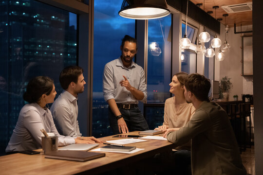 Multiracial businesspeople work in office late hours, discuss financial project at team meeting. Diverse multiethnic colleagues coworkers busy cooperate in boardroom at night. Deadline concept.