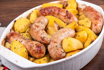 Baked golden pieces of potatoes with turkey sausages in a white ceramic baking dish on a wooden table, close-up