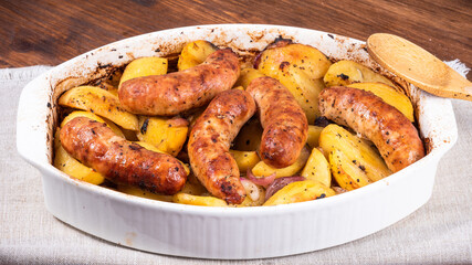 Baked golden pieces of potatoes with turkey sausages in a white ceramic baking dish on a wooden table, close-up