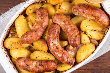 Baked golden pieces of potatoes with turkey sausages in a white ceramic baking dish on a wooden table, top view, close-up