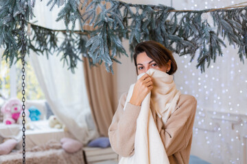 Warm portrait of happy woman in sweater, jeans and white scarf  at home in bedroom,  christmas lights on background. 